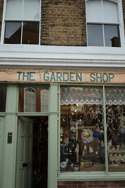 The image shows the exterior of a garden shop called 'The Garden Shop' situated on the ground floor of a brick building. The shop has light green painted wooden frames around large display windows fil